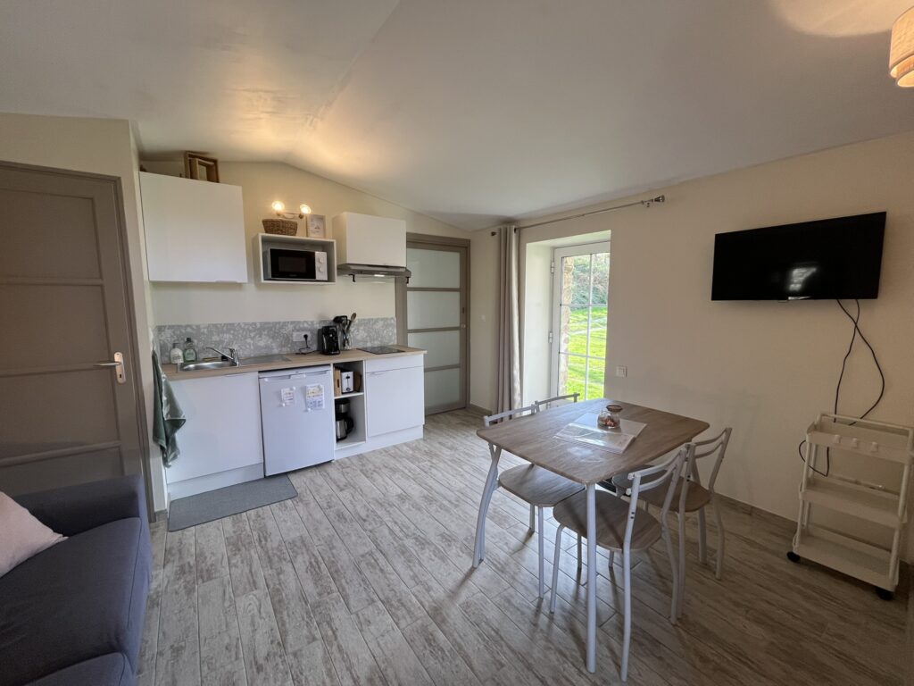 Cuisine et salle à manger compactes et modernes avec des armoires blanches, une table en bois avec quatre chaises, une télévision murale et une porte vitrée ouvrant sur un espace extérieur herbeux.