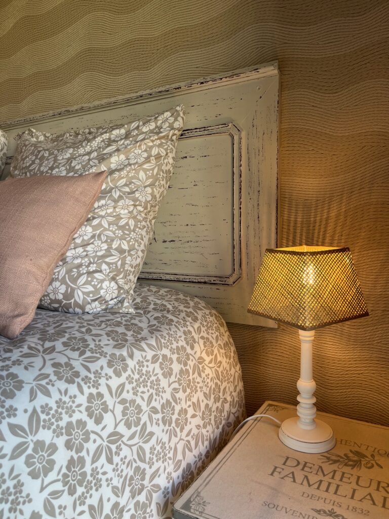 Un lit avec une literie florale et un oreiller rose se trouve à côté d'une table de nuit avec une lampe allumée et une boîte décorative, contre un mur beige texturé.