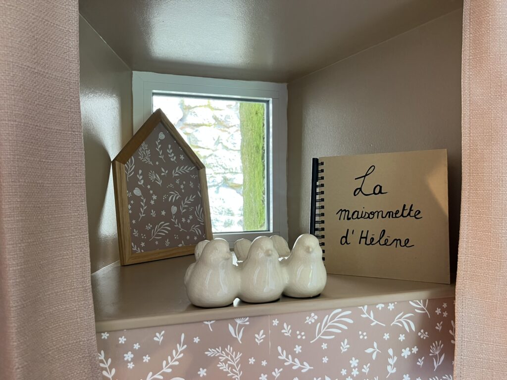 Une petite étagère contient un décor en forme de maison, un cahier intitulé La Maisonnette d'Hélène, et quatre figurines d'oiseaux en céramique blanche devant une fenêtre.