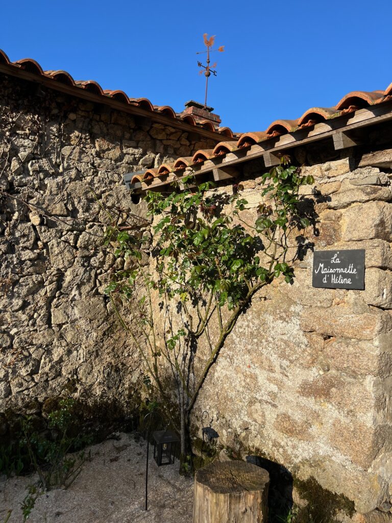 Bâtiment en pierre avec un toit en tuiles, un petit arbre devant, et un panneau indiquant La Maisonnette d'Hélène sur le mur, sous un ciel bleu clair.