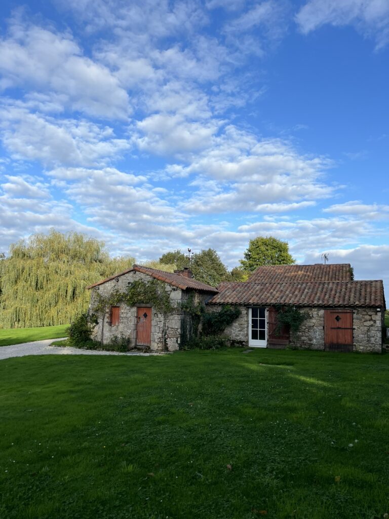 Une maison en pierre avec un toit de tuiles rouges et des volets rouges est posée sur une pelouse sous un ciel bleu partiellement nuageux.