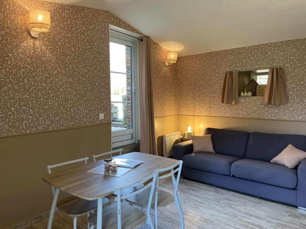 Un petit salon avec un papier peint à motifs, un canapé gris, une table à manger en bois avec des chaises, et une fenêtre avec des rideaux qui laissent entrer la lumière naturelle.