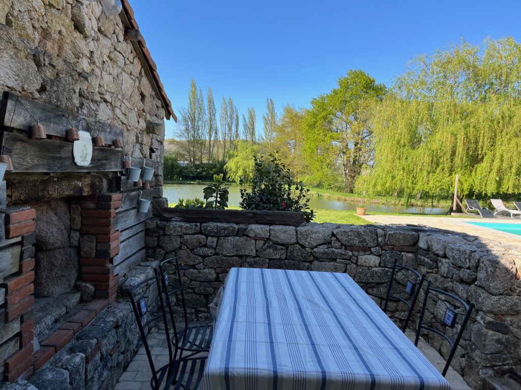 Patio en pierre avec une nappe rayée sur une table et des chaises noires, donnant sur un étang avec des arbres et une piscine par une journée ensoleillée.