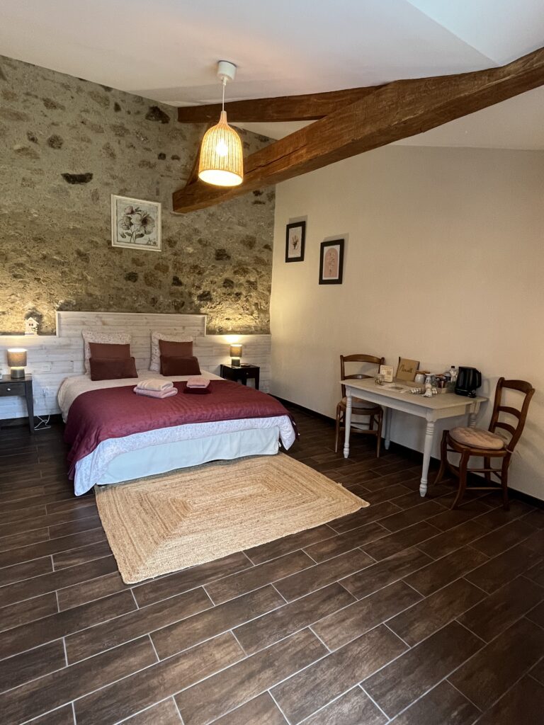 Une chambre confortable avec un lit double, deux tables de chevet, un bureau avec des chaises, des poutres en bois apparentes et un mur d'accent en pierre. Les serviettes sont pliées sur le lit.