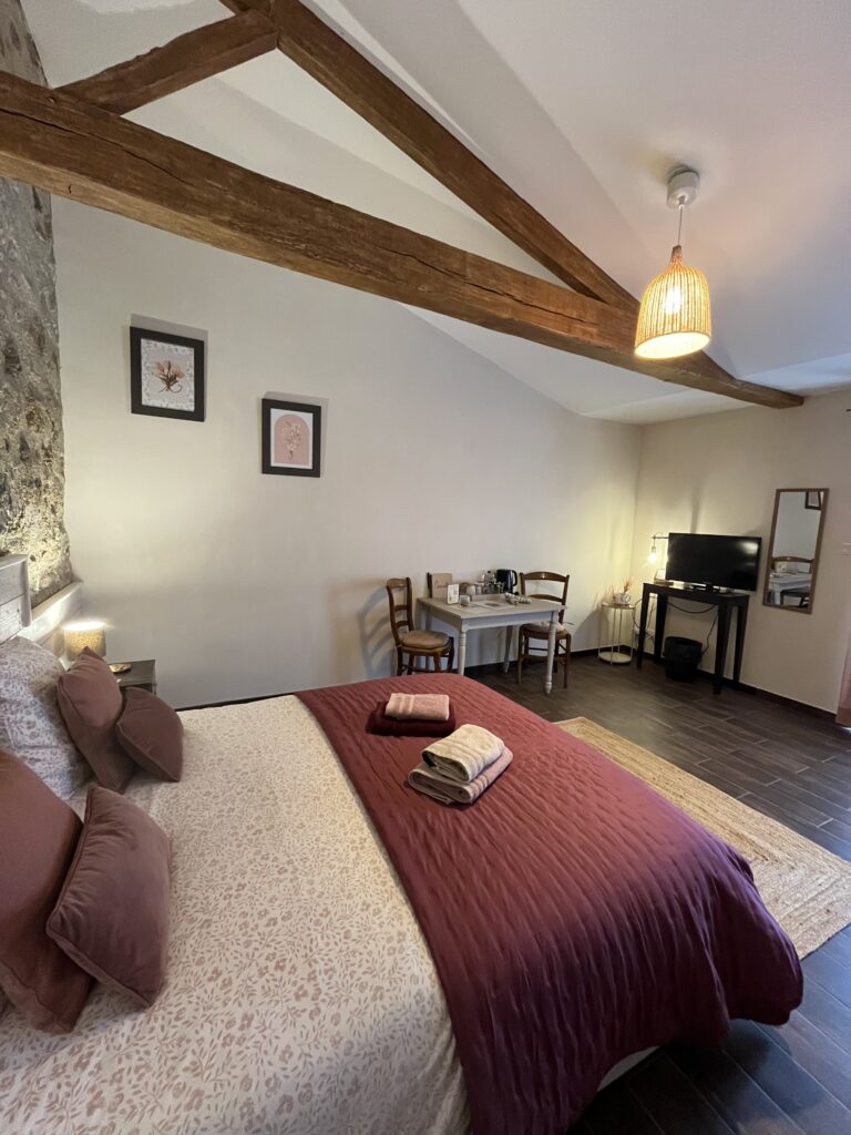 Une chambre confortable avec un lit double, deux chaises sur un bureau, une télévision, des poutres en bois apparentes, des serviettes pliées sur le lit et des œuvres d'art encadrées au mur.