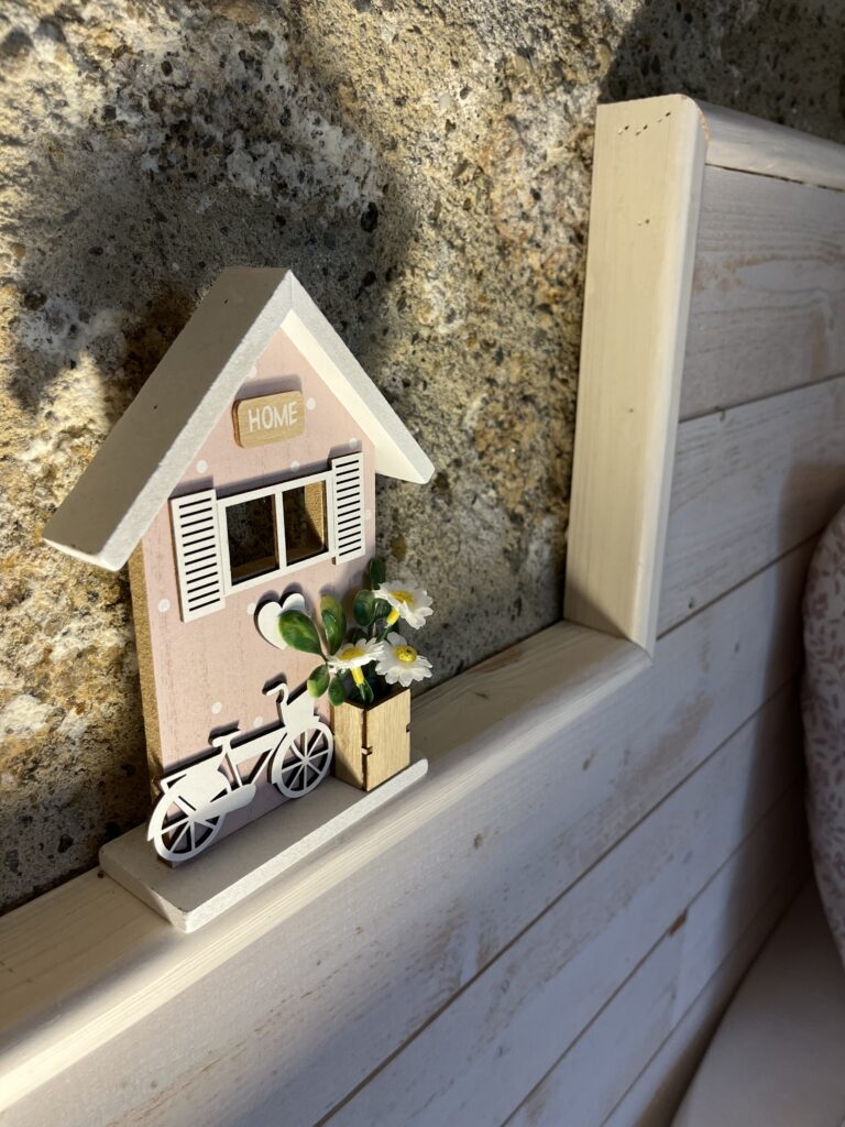 Une petite maison décorative avec un vélo et un pot de fleurs est posée sur une étagère en bois à côté d'un mur en pierre, éclairée par la lumière du soleil.