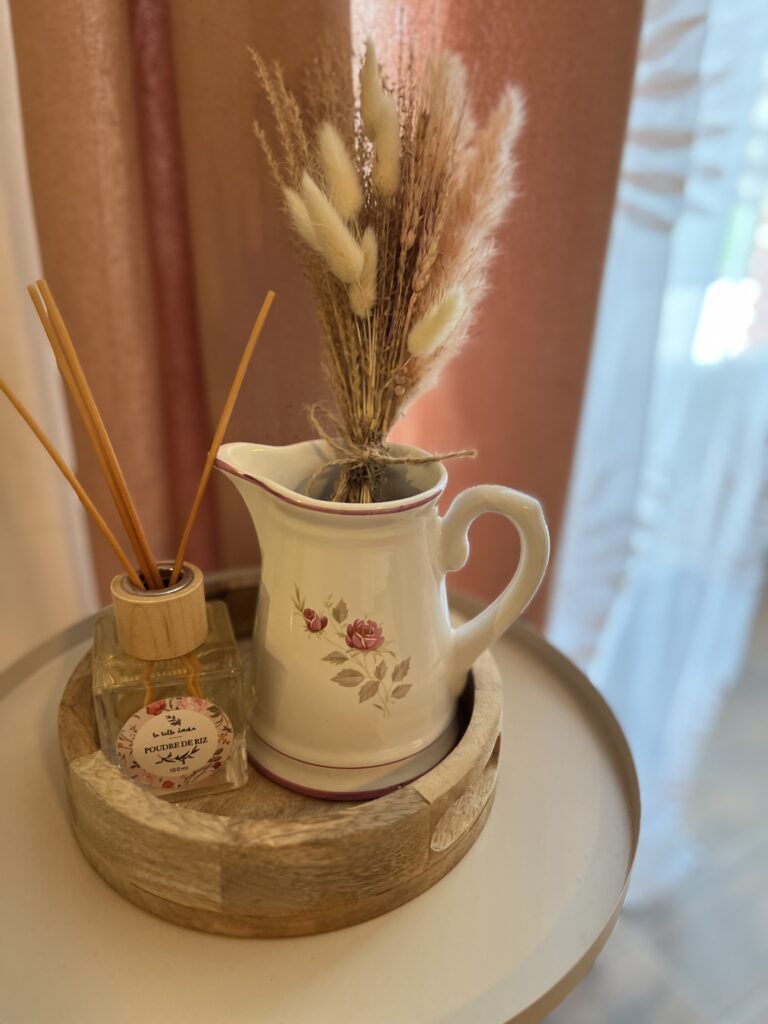 Un plateau en bois contient un pichet en céramique avec des fleurs séchées et un flacon de diffuseur à roseaux sur une table ronde à côté de rideaux transparents.