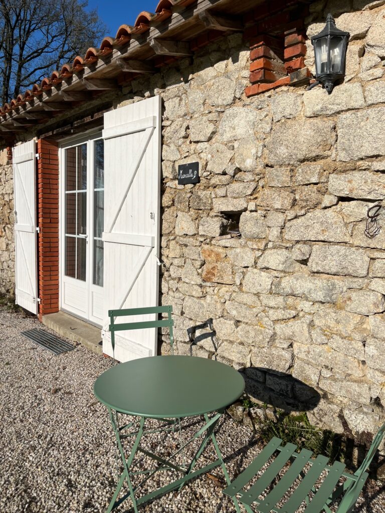 Une petite table en métal vert avec deux chaises se trouve à l'extérieur d'un bâtiment en pierre avec des volets blancs, des portes en verre et une lanterne murale à la lumière du soleil.