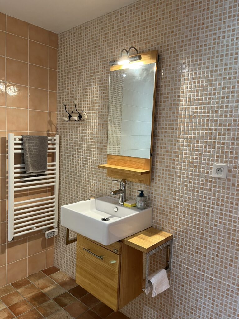 Une salle de bains avec un lavabo mural, un meuble en bois, un miroir avec lumière, un porte-serviettes avec une serviette grise et des murs en carreaux de mosaïque beige.
