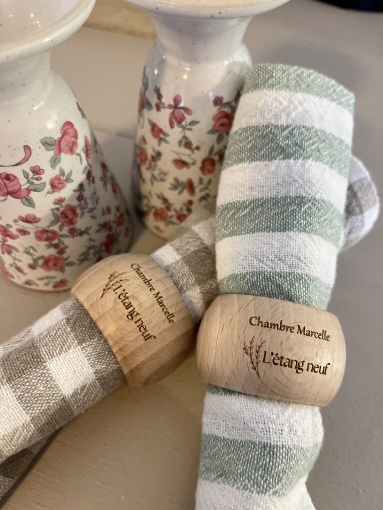 Deux serviettes, l'une à rayures et l'autre à carreaux, chacune retenue par un rond de serviette en bois gravé Chambre Marcelle Létang neuf, sont placées à côté de deux vases en céramique à motifs floraux.