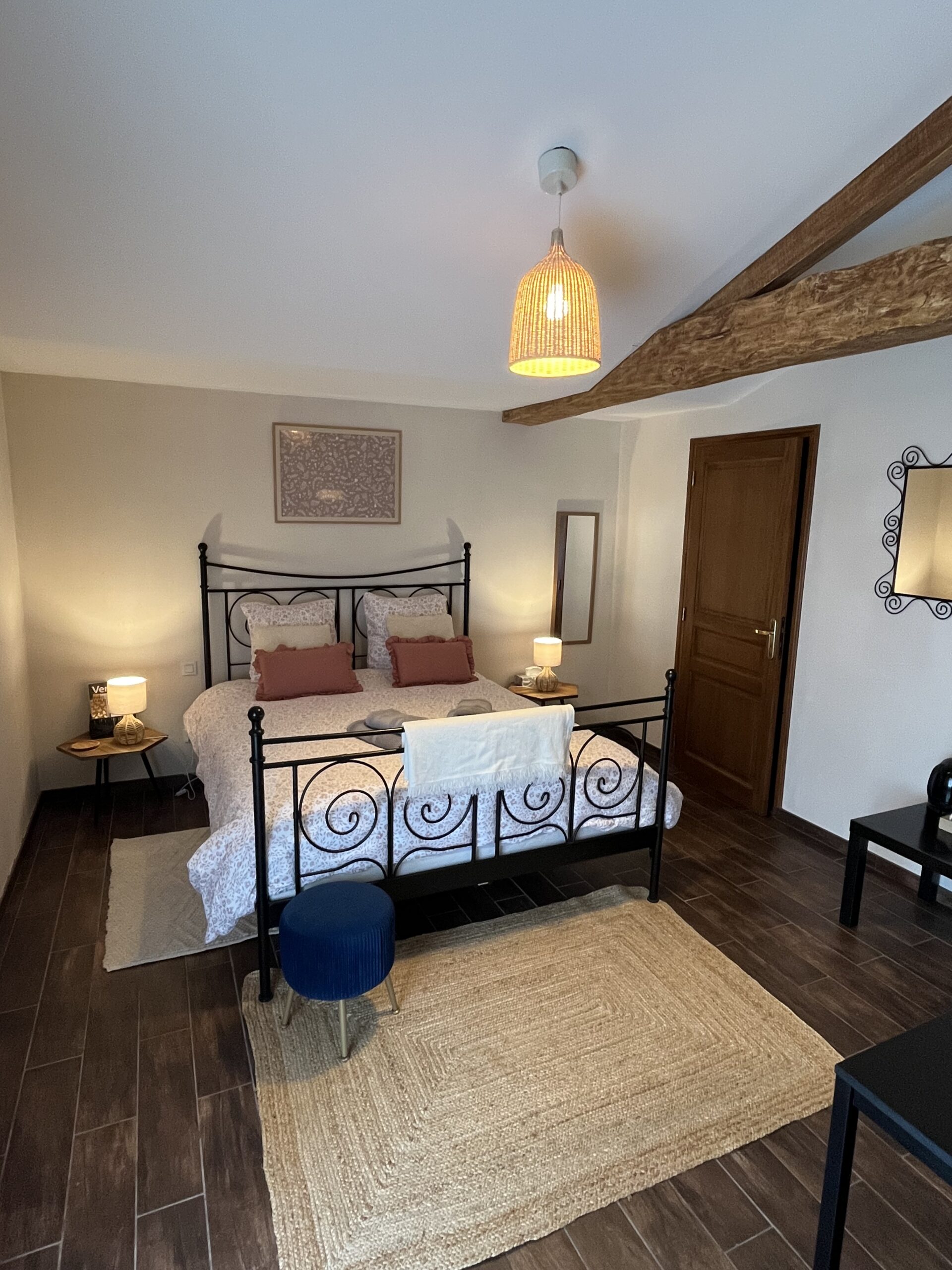 Un lit double bien fait avec une literie à motifs, deux coussins roses et deux tables de chevet avec des lampes dans une chambre confortable avec des poutres en bois et un tapis tissé.