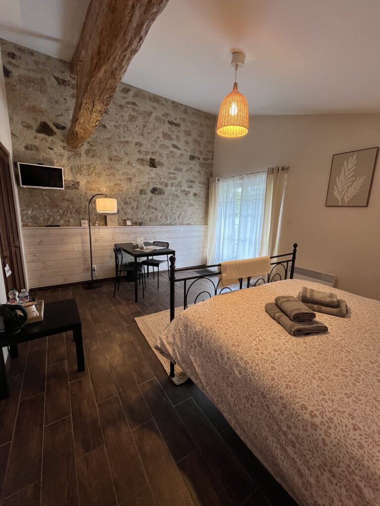 Chambre confortable avec un lit double, des serviettes pliées, un sol en bois, un mur en pierres apparentes, un petit bureau, une télévision murale et une fenêtre avec des rideaux transparents laissant entrer la lumière naturelle.