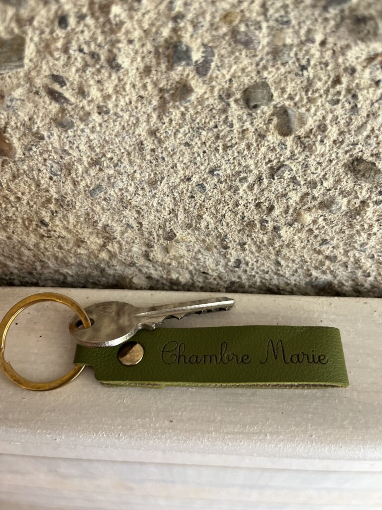 Une clé avec un porte-clés en or attaché à une étiquette en cuir vert portant l'inscription Chambre Marie, placée sur une surface en bois clair contre un mur en pierre.