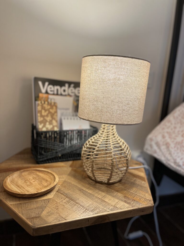 Une lampe de table tressée est allumée sur une table de nuit en bois, à côté de deux sous-verres en bois et d'un panier noir contenant des magazines et des papiers.