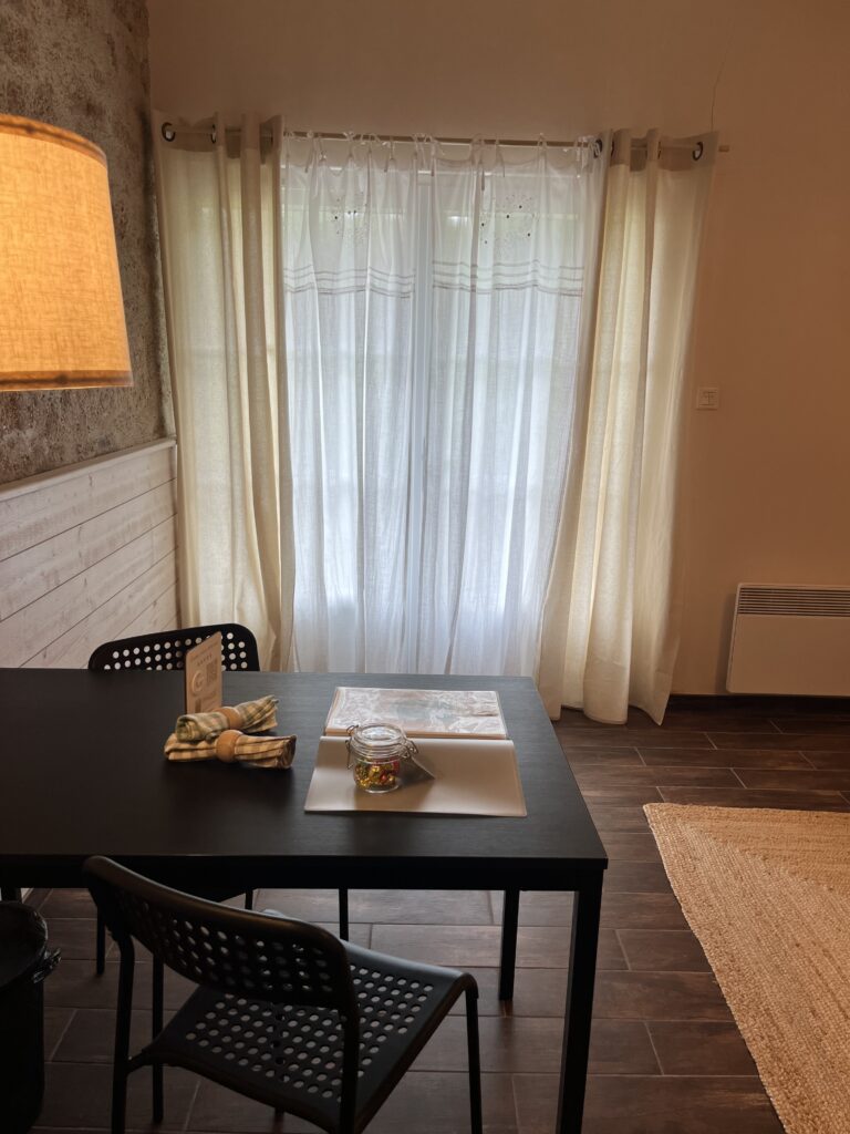 Une table noire avec un pot en verre, un set de table, une serviette pliée et des papiers se trouve devant des rideaux blancs transparents dans une pièce à l'éclairage tamisé et au sol sombre.