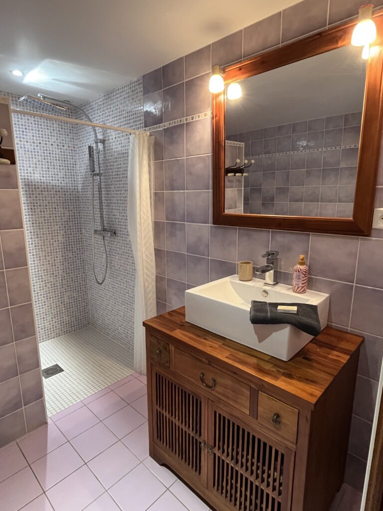 Salle de bains avec des murs carrelés en violet, une douche à l'italienne avec un rideau, et un meuble-lavabo en bois avec un lavabo carré, un miroir et des articles de toilette.