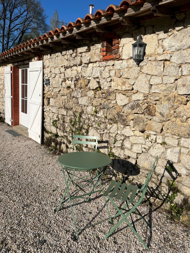 Une petite table en métal vert et deux chaises sont placées sur du gravier à l'extérieur d'un bâtiment rustique en pierre avec des doubles portes blanches et une lanterne murale.
