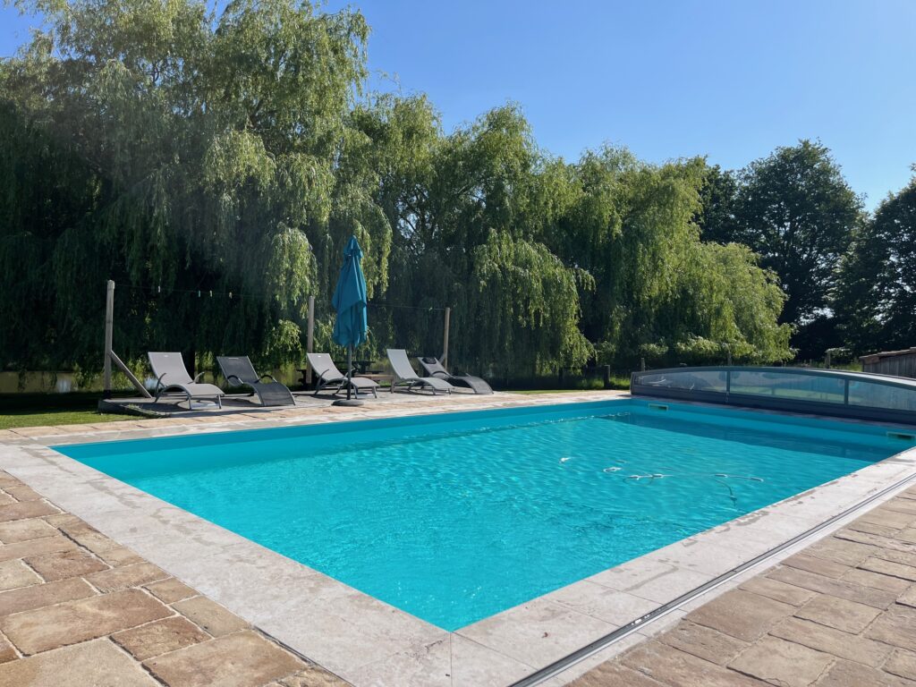 Piscine extérieure rectangulaire avec une eau bleue claire, entourée de chaises longues, d'un parasol et d'arbres en arrière-plan sous un ciel dégagé.