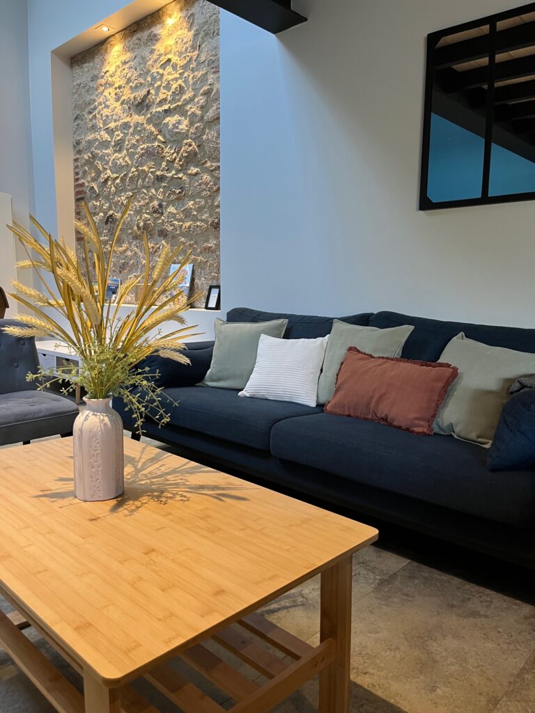 Un salon moderne avec un canapé bleu foncé, des coussins assortis, une table basse en bois avec un vase de plantes séchées et un mur d'accentuation en pierre avec des spots.