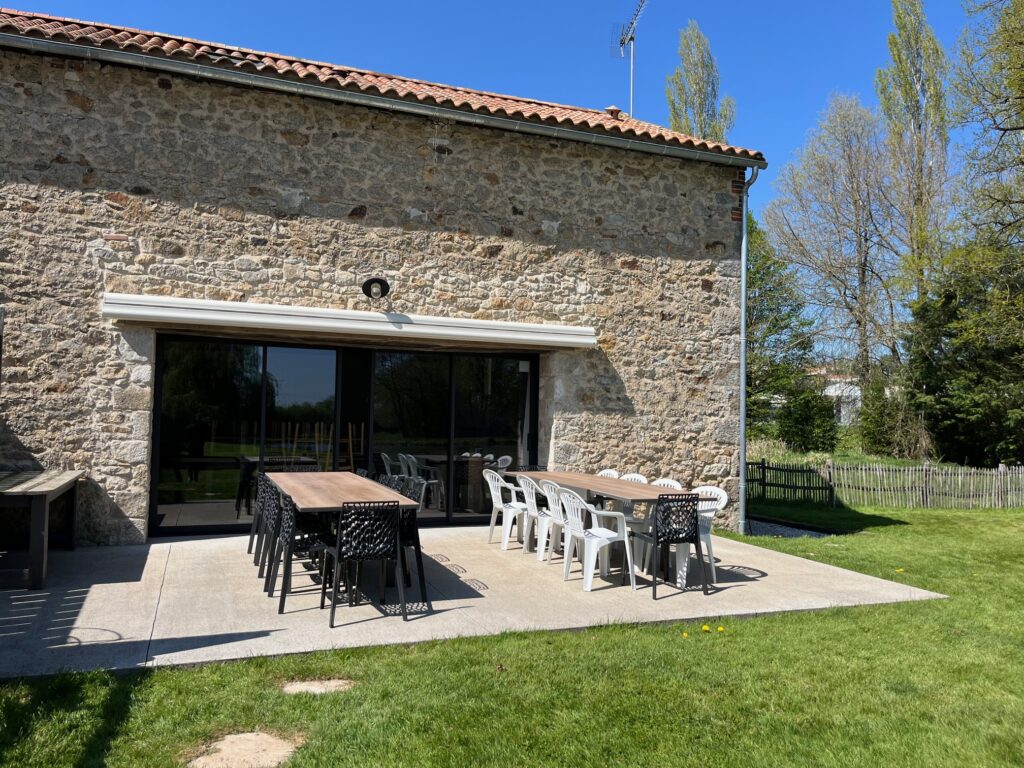 Patio extérieur avec deux longues tables et des chaises mixtes noires et blanches, adossé à un bâtiment en pierre avec de grandes portes coulissantes en verre, lors d'une journée ensoleillée.
