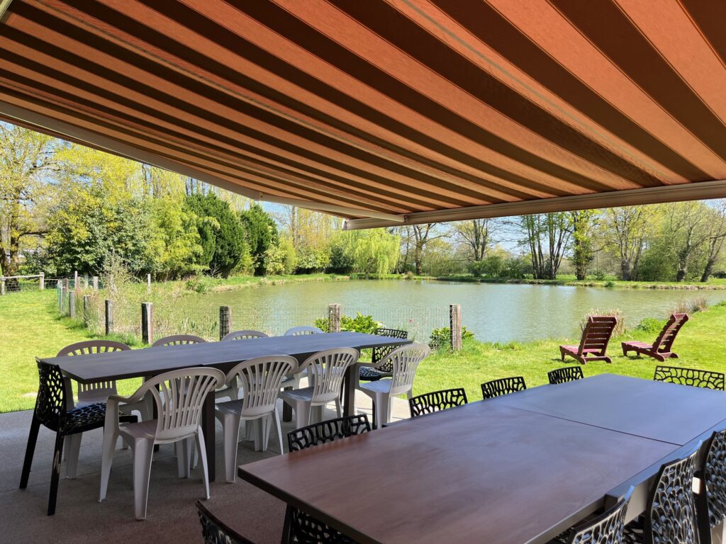 Le patio extérieur couvert avec deux tables et des chaises mixtes donne sur un étang, une zone herbeuse et des arbres. Deux chaises longues en bois se trouvent sur la pelouse, près de l'eau.