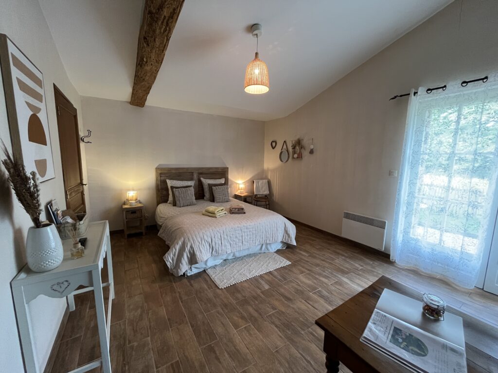 Un lit bien fait et des tables de chevet dans une chambre spacieuse et lumineuse, avec du parquet, un décor minimaliste et une fenêtre aux rideaux diaphanes qui laissent entrer la lumière naturelle.