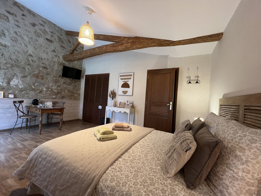 Chambre confortable avec un lit double, une literie neutre, des poutres en bois, un mur d'accent en pierre, un petit bureau, des œuvres d'art murales et une télévision accrochée au mur. Les serviettes sont soigneusement placées sur le lit.