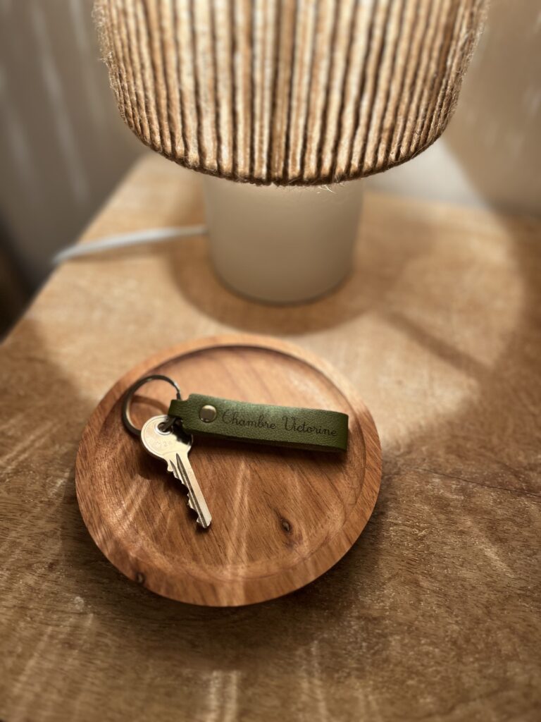 Un trousseau de clés avec un porte-clés vert étiqueté Chambre Udeme repose sur un plat en bois à côté d'une lampe sur une surface en bois.