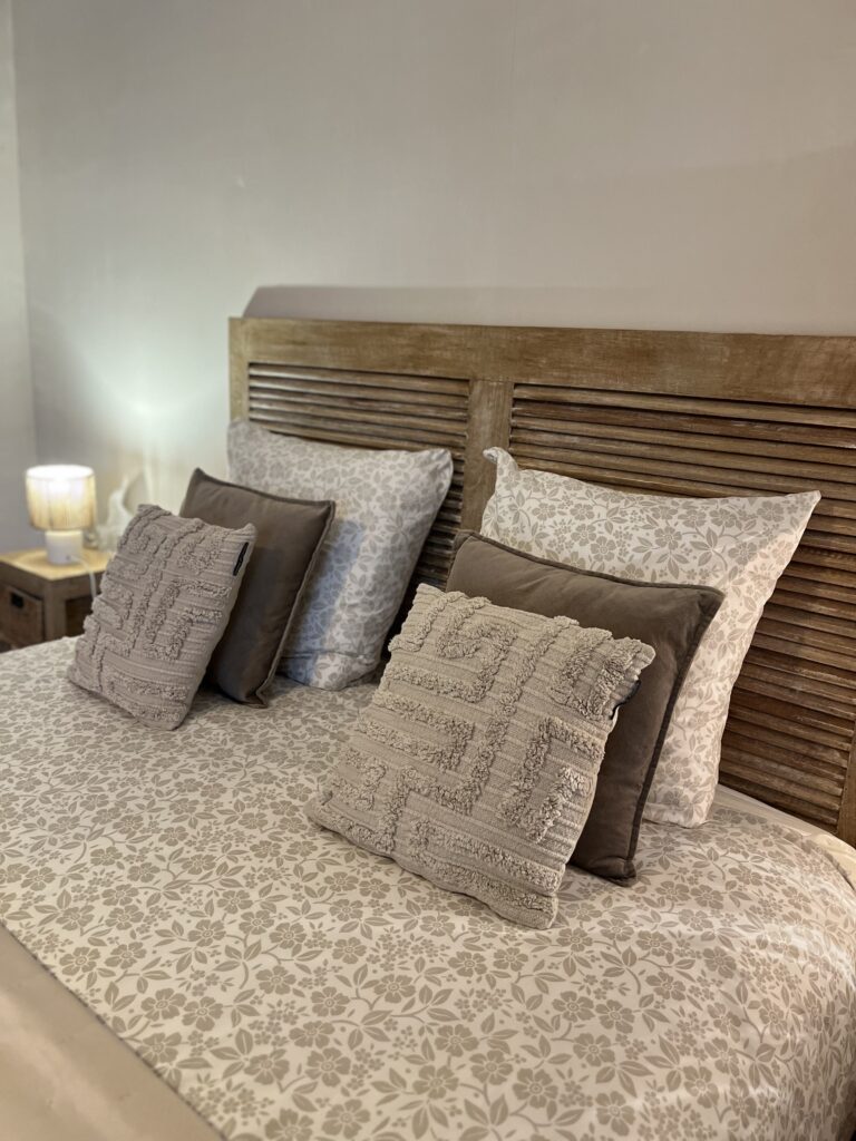 Un lit avec une tête de lit en bois est garni d'une literie à motifs floraux, de deux coussins d'appoint bruns et de deux coussins jetés texturés beiges. Une lampe allumée est posée sur la table de chevet.