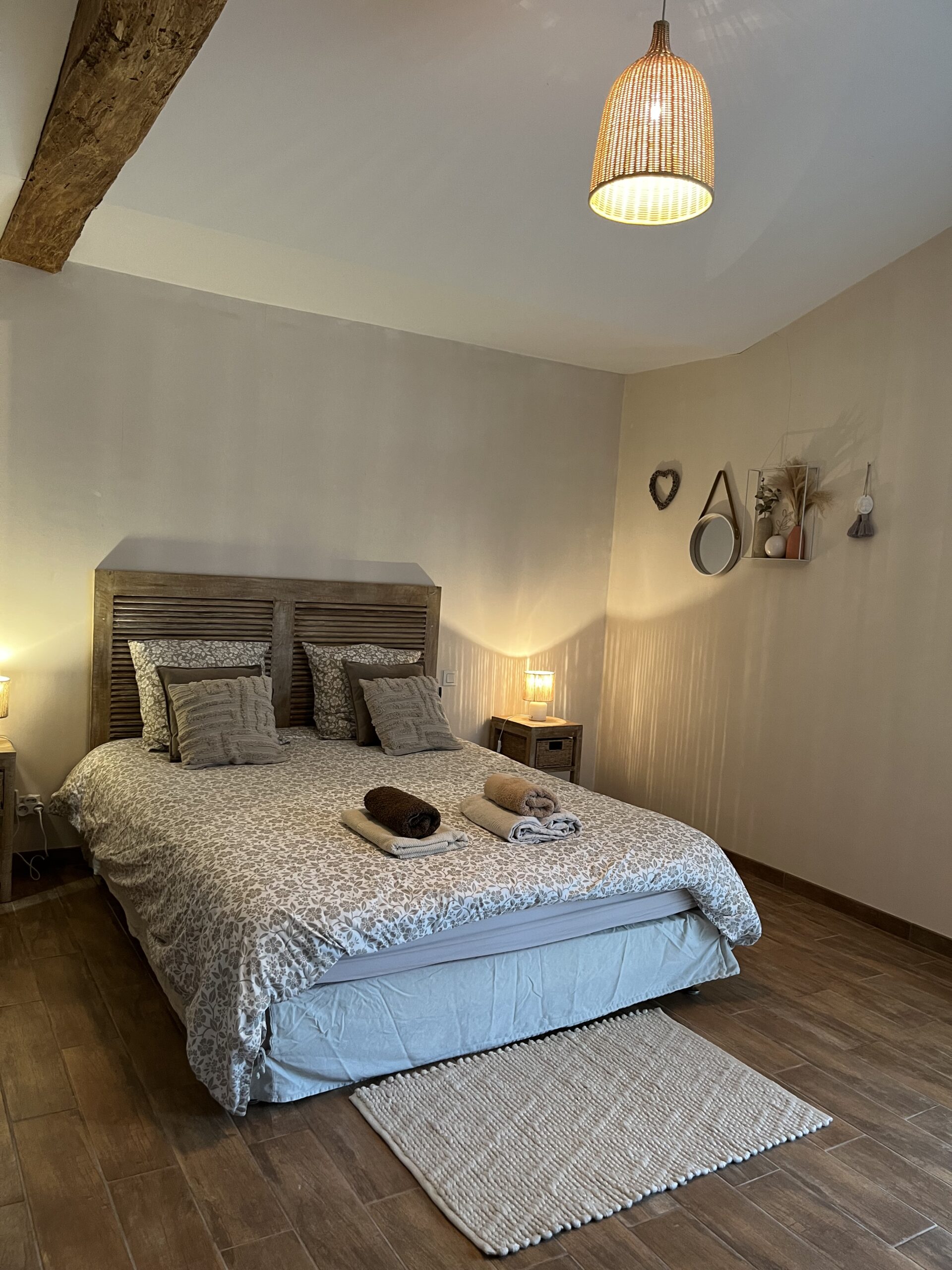 Une chambre confortable avec un lit double, deux tables de chevet avec lampes, des serviettes pliées sur le lit et des décorations murales. Le sol est en bois et une lampe suspendue est accrochée au plafond.