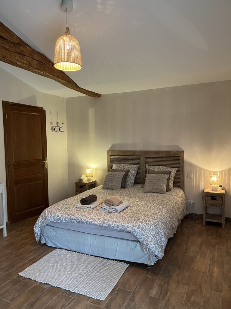 Un lit double bien fait avec des serviettes pliées sur le dessus, des tables de chevet avec des lampes, un plancher en bois, une porte et un luminaire suspendu dans une chambre à coucher confortable.