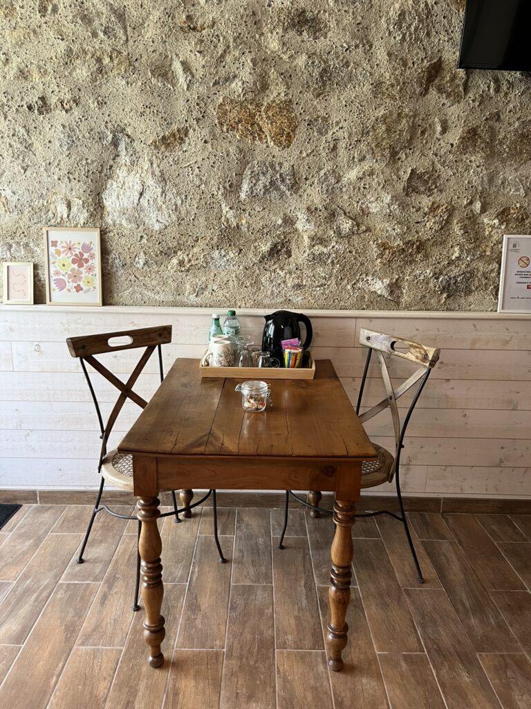 Une table en bois avec deux chaises, un sucrier en verre et un plateau contenant des bocaux et une théière, posés contre un mur de pierre rustique dans un petit café.