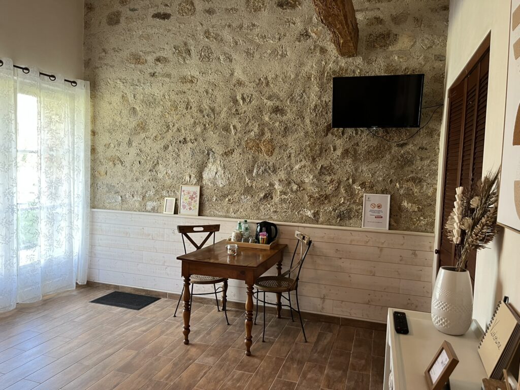 Une petite table en bois avec deux chaises est posée contre un mur de pierre dans une pièce lumineuse avec une télévision montée, des tableaux encadrés et des rideaux diaphanes près d'une fenêtre.