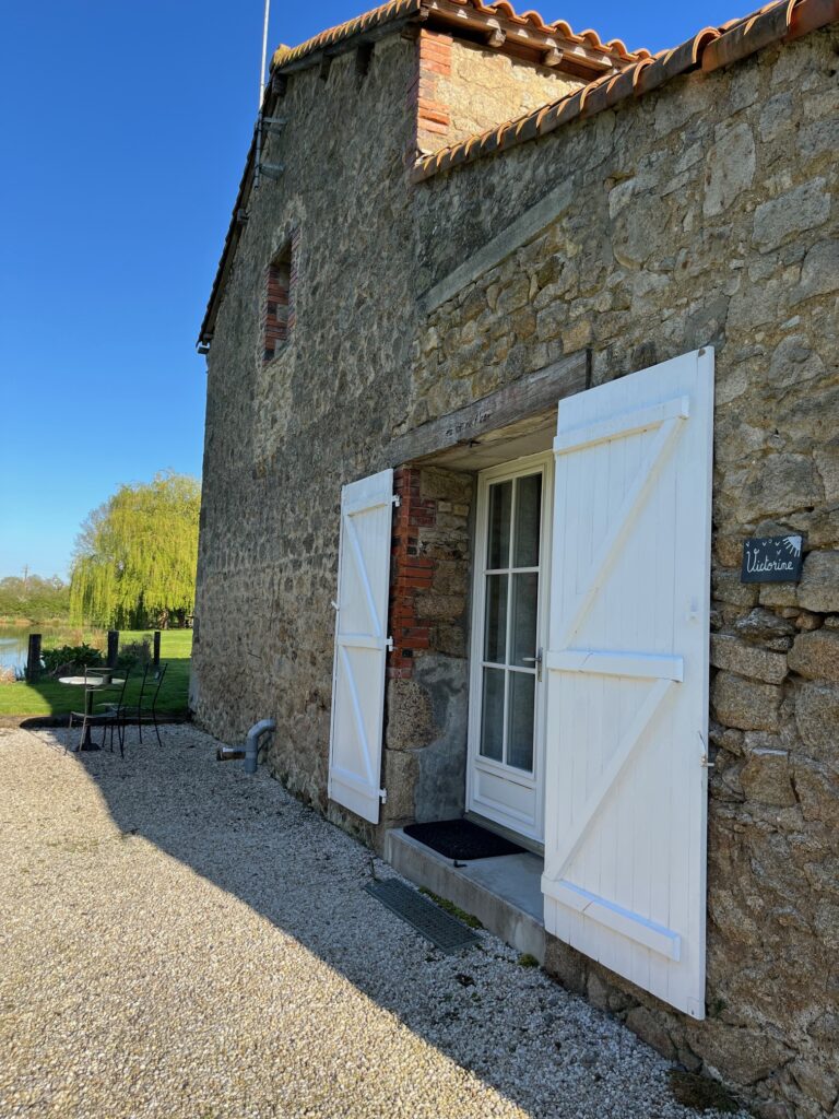 Bâtiment en pierre avec doubles portes blanches ouvrant sur un chemin de gravier ; une petite table et des chaises sont visibles sur une pelouse sous un ciel bleu clair.