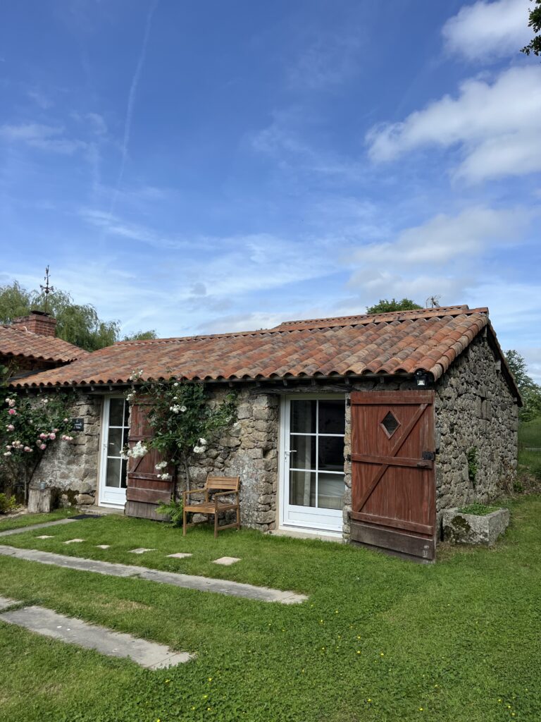 Une petite maison en pierre avec un toit en tuiles rouges, des portes vitrées à cadre blanc, des volets en bois et un banc en bois, le tout sur une pelouse sous un ciel bleu.
