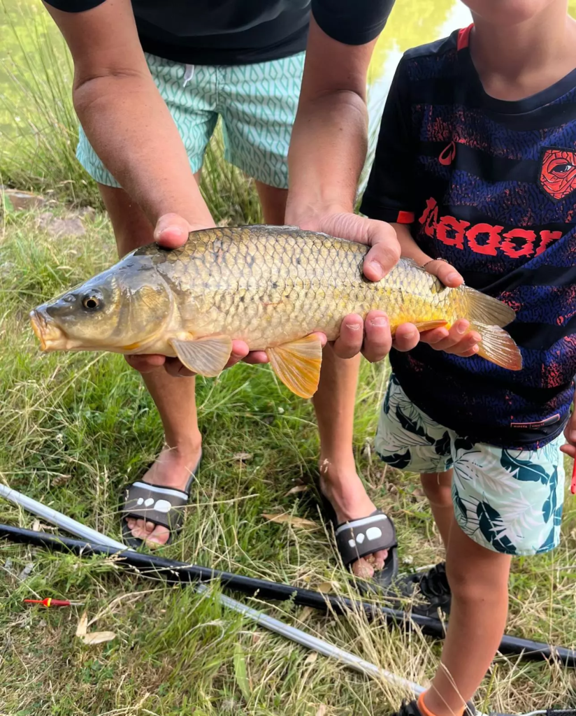 Deux personnes, à l'extérieur des Gîtes du Puy du Fou, tiennent fièrement à l'horizontale, à deux mains, un poisson fraîchement pêché ; les cannes à pêche et l'herbe viennent rehausser le cadre pittoresque de la Vendée.